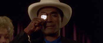 Movie still from “Wild at Heart” (1990), directed by David Lynch – A man wearing a white hat holding a coin in front of his face; Close Up shot, Over the shoulder angle