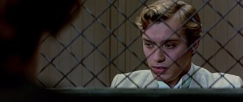 Movie still from “Wilde” (1997), directed by Brian Gilbert – A young man sitting in front of a chain link fence; Close Up shot, Over the shoulder angle