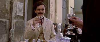 Movie still from “Wilde” (1997), directed by Brian Gilbert – A man sitting at a table holding a wine glass; Medium shot, Over the shoulder angle