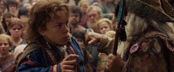 Movie still from “Willow” (1988), directed by Ron Howard – A young boy is holding a stick in front of a group of people; Close Up shot, Over the shoulder angle
