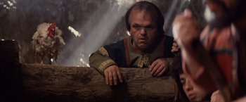 Movie still from “Willow” (1988), directed by Ron Howard – A man in a medieval outfit leaning on a wooden fence; Medium shot, Low angle