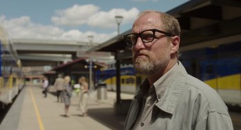 Movie still from “Wilson” (2017), directed by Craig Johnson – A man with a goatee and glasses is standing in front of a train station; Medium shot, Over the shoulder angle