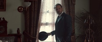Movie still from “Winchester” (2018), directed by Michael Spierig – A man in a suit and tie holding a hat; Medium shot, Low angle