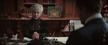 Movie still from “Winchester” (2018), directed by Michael Spierig – An older woman sitting at a table in front of a painting; Medium shot, Over the shoulder angle