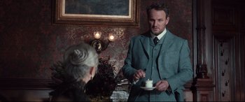 Movie still from “Winchester” (2018), directed by Michael Spierig – A man in a suit and tie is holding a cup; Medium shot, Over the shoulder angle