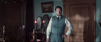 Movie still from “Winchester” (2018), directed by Michael Spierig – A man standing in a room wearing a suit and tie; Medium shot, Low angle