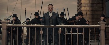 Movie still from “Winter's Tale” (2014), directed by Akiva Goldsman – A man in a suit and tie standing in front of a fence; Medium shot, Low angle