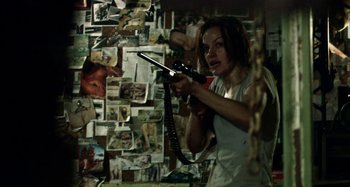 Movie still from “Wolf Creek” (2005), directed by Greg McLean – A woman holding a gun in front of a wall covered in pictures; Medium shot, High angle