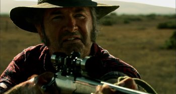 Movie still from “Wolf Creek” (2005), directed by Greg McLean – A man with a rifle in a field; Close Up shot, Over the shoulder angle