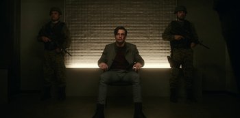 Movie still from “Another Life” (2019), created by Aaron Martin – A man sitting on a chair in front of two soldiers; Wide shot, Low angle
