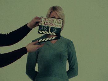 Movie still from “Women Reply” (1975), directed by Agnès Varda – A woman holding a clapperboard over her face; Medium shot, Low angle