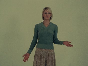 Movie still from “Women Reply” (1975), directed by Agnès Varda – A woman standing in front of a white wall with her arms outstreched; Medium shot, Low angle