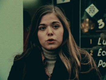 Movie still from “Women Reply” (1975), directed by Agnès Varda – A woman with long brown hair wearing a black coat; Close Up shot, Low angle
