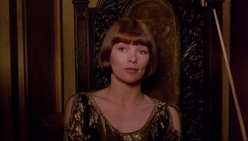 Movie still from “Women in Love” (1969), directed by Ken Russell – A woman in a black and gold dress sitting in front of a wooden chair; Close Up shot, Over the shoulder angle