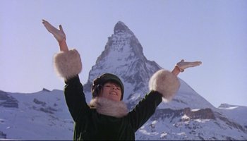 Movie still from “Women in Love” (1969), directed by Ken Russell – A woman in a green coat and white gloves in front of a snow covered mountain; Medium shot, Low angle