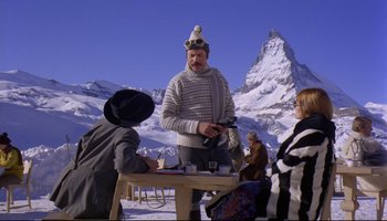 Movie still from “Women in Love” (1969), directed by Ken Russell – A man wearing a hat standing at a wooden table in the snow; Wide shot, Low angle