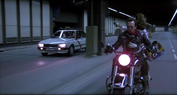 Movie still from “Women on the Verge of a Nervous Breakdown” (1988), directed by Pedro Almodóvar – A man riding a motorcycle down a street next to a car; Wide shot, Low angle