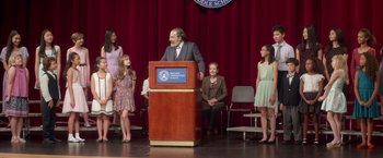 Movie still from “Wonder” (2017), directed by Stephen Chbosky – A man giving a speech to a group of people on a stage; Wide shot, Low angle
