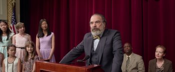 Movie still from “Wonder” (2017), directed by Stephen Chbosky – A man in a suit and a bow tie standing at a podium; Medium shot, Over the shoulder angle
