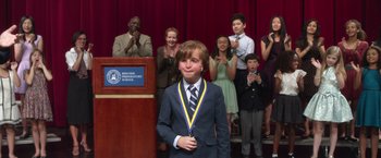 Movie still from “Wonder” (2017), directed by Stephen Chbosky – A young boy wearing a medal standing in front of an audience; Medium shot, Over the shoulder angle