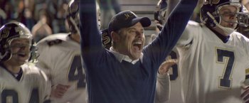 Movie still from “Woodlawn” (2015), directed by Jon Erwin – A man in a blue sweater and a hat is holding his hands in the air; Medium shot, Low angle