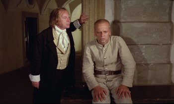 Movie still from “Woyzeck” (1979), directed by Werner Herzog – Two men dressed in historical clothing , one of them sitting on the ground; Medium shot, Low angle