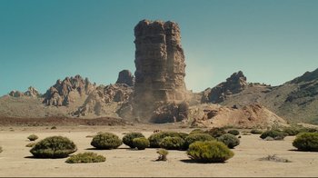Movie still from “Wrath of the Titans” (2012), directed by Jonathan Liebesman – A tall rock formation in the desert with a bunch of bushes in front of it; Extreme Wide shot, High angle