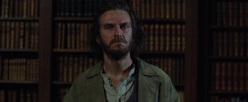 Movie still from “Apostle” (2018), directed by Gareth Evans – A man with long hair standing in front of a book shelf; Close Up shot, Low angle