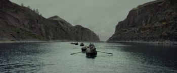 Movie still from “Apostle” (2018), directed by Gareth Evans – A group of people in a row boat on a body of water; Extreme Wide shot, High angle