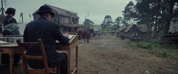 Movie still from “Apostle” (2018), directed by Gareth Evans – A man sitting at a desk in a dirt field; Wide shot, Over the shoulder angle