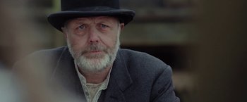 Movie still from “Apostle” (2018), directed by Gareth Evans – An older man wearing a black hat and a suit; Close Up shot, Over the shoulder angle