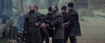 Movie still from “Apostle” (2018), directed by Gareth Evans – A group of men in coats and hats standing next to each other on a field; Medium shot, Over the shoulder angle