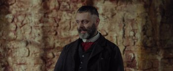 Movie still from “Apostle” (2018), directed by Gareth Evans – A man with a gray beard wearing a black suit; Close Up shot, Low angle