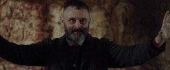 Movie still from “Apostle” (2018), directed by Gareth Evans – A man with a beard and a suit and tie; Close Up shot, Low angle