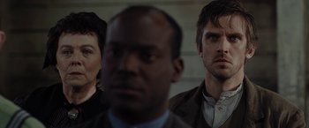 Movie still from “Apostle” (2018), directed by Gareth Evans – A man and a woman standing next to each other in front of another man; Close Up shot, Over the shoulder angle