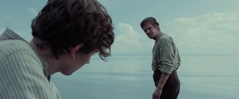 Movie still from “Apostle” (2018), directed by Gareth Evans – A man standing next to another man on a beach; Medium shot, Over the shoulder angle