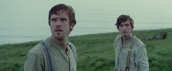 Movie still from “Apostle” (2018), directed by Gareth Evans – A man and a woman standing next to each other on top of a grass covered hill; Close Up shot, Low angle