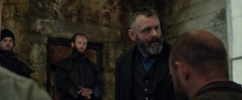 Movie still from “Apostle” (2018), directed by Gareth Evans – A man with a long beard and a mustache wearing a suit; Medium shot, Over the shoulder angle