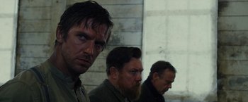 Movie still from “Apostle” (2018), directed by Gareth Evans – A group of men standing next to each other in front of a brick wall; Close Up shot, Over the shoulder angle