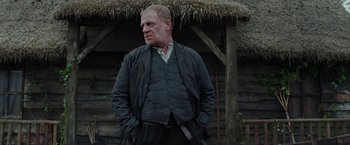 Movie still from “Apostle” (2018), directed by Gareth Evans – A man standing in front of a wooden house; Medium shot, Low angle