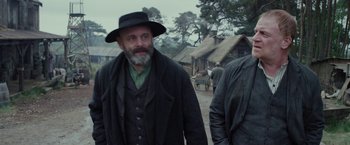 Movie still from “Apostle” (2018), directed by Gareth Evans – A man with a long beard wearing a black hat; Medium shot, Low angle