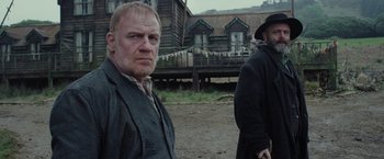 Movie still from “Apostle” (2018), directed by Gareth Evans – Two men standing in front of an old house; Medium shot, Low angle