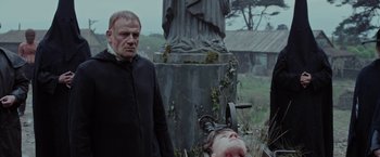 Movie still from “Apostle” (2018), directed by Gareth Evans – A man standing next to a statue of a woman laying on the ground; Medium shot, Low angle