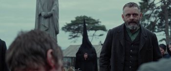 Movie still from “Apostle” (2018), directed by Gareth Evans – A man in a black suit and a black hat; Medium shot, Over the shoulder angle