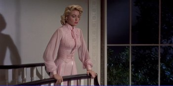 Movie still from “Written on the Wind” (1956), directed by Douglas Sirk – A woman in a pink dress leaning on a railing in front of a window; Medium shot, Low angle