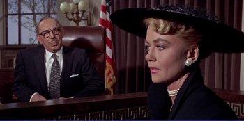 Movie still from “Written on the Wind” (1956), directed by Douglas Sirk – A woman wearing a hat sitting in front of a judge; Medium shot, Over the shoulder angle
