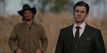 Movie still from “Yellowstone” (2018), created by John Linson – A man in a suit standing next to another man in a suit; Medium shot, Over the shoulder angle