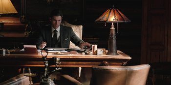 Movie still from “Yellowstone” (2018), created by John Linson – A man sitting at a table with a lamp on top of a table; Medium shot, Over the shoulder angle