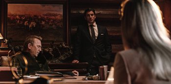 Movie still from “Yellowstone” (2018), created by John Linson – A man standing in front of a table with other people; Medium shot, Over the shoulder angle