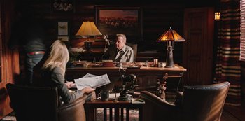 Movie still from “Yellowstone” (2018), created by John Linson – An older man sitting at a desk in front of a woman; Wide shot, High angle